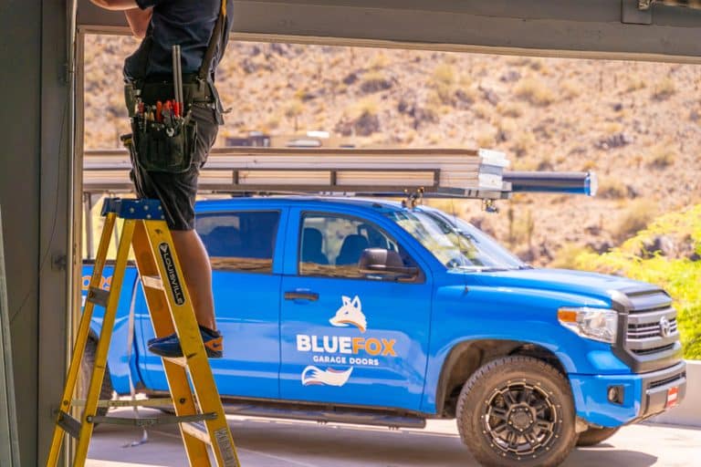 man on ladder fixing garage door with blue fox truck in backround